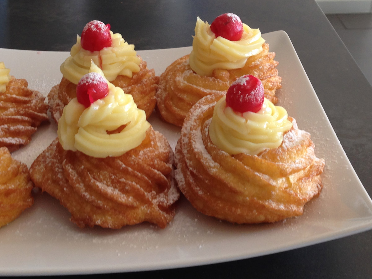 St Joseph fried zeppole on a white rectangle dish with on top pastry cream and a candied cherry.