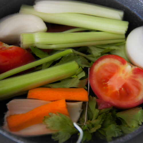 Pot filled with fresh vegetables: tomatoes, carrots, celery, onion, parsley, submerged in water to make homemade brodo vegetale.