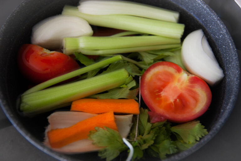 Pot filled with fresh vegetables: tomatoes, carrots, celery, onion, parsley, submerged in water to make homemade brodo vegetale.