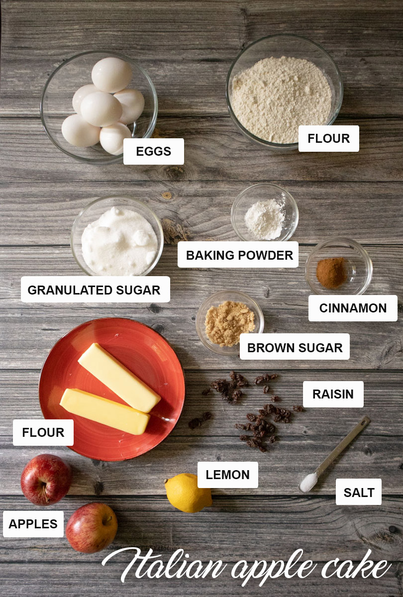 Ingredients for Italian apple cake arranged on a table, including fresh apples, eggs, flour, sugar, butter, and cinnamon.