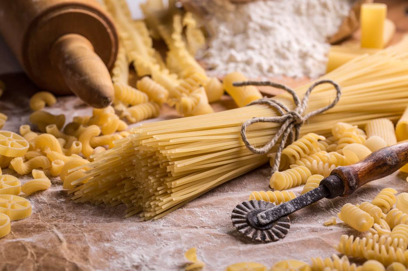 Dry spaghetti and assorted pasta shapes on a floured surface with traditional pasta-making tools. Dry spaghetti and assorted pasta shapes on a floured surface with traditional pasta-making tools.