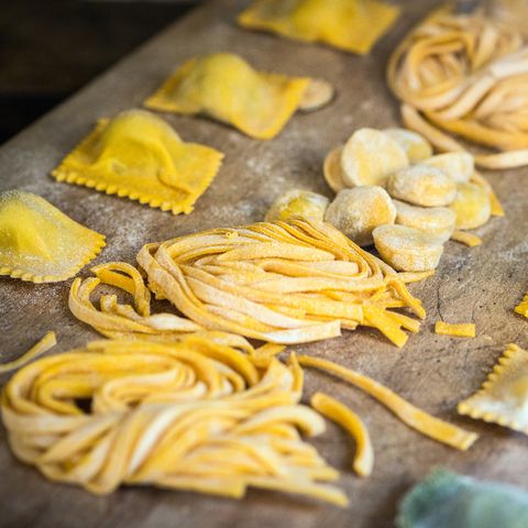 Assorted homemade fresh pasta shapes arranged on a wooden cutting board, including tagliatelle, fettuccine, ravioli.
