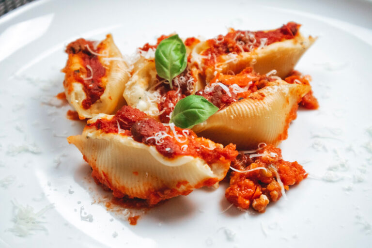 Close-up of baked jumbo pasta shells filled with ricotta and cooked ham, topped with meat ragù and melted cheese, served on a white plate. Garnished with two basil leaves.